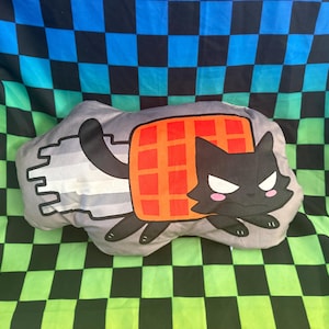 May include: A gray plush pillow shaped like a cartoon cat with a waffle body. The cat is black with white eyes and pink cheeks. The waffle is orange with a grid pattern. The pillow is on a checkered background.