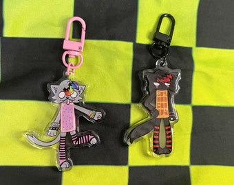 May include: Two cartoon cat keychains. One is gray with rainbow hair, a pink dress, and black and pink striped socks. The other is black with red hair and a waffle design on its torso. Both have metal clasps.
