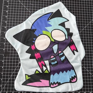 May include: A light blue plush pillow featuring a cartoon monster design. The monster has large white eyes, a purple body, and accents of black, blue, pink, and green. The pillow is on a black cutting mat.