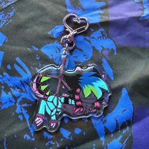 Puede incluir: Un llavero acrílico transparente con un personaje de dibujos animados con cabello azul y verde y ropa turquesa y rosa. El llavero tiene un cierre plateado en forma de corazón.