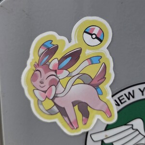 Puede incluir: Una pegatina con un personaje Sylveon rosa con un borde amarillo. El diseño incluye una Poké Ball con los colores del orgullo trans. Sylveon tiene una pajarita y cintas que fluyen, con una expresión feliz.