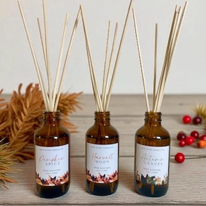 May include: Three brown glass reed diffusers with wooden reeds. Each bottle has a label with a fall-themed name: Pumpkin Spice, Harvest Moon, and Autumn Leaves. The bottles are arranged on a light wood surface.