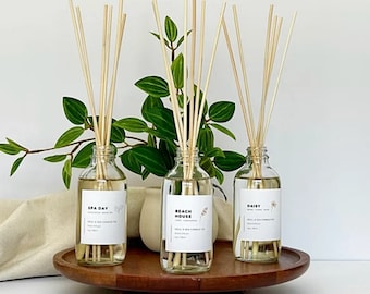 Puede incluir: Tres botellas de vidrio con difusores de caña y etiquetas que dicen "Spa Day", "Beach House" y "Daisy". Las botellas están sobre una bandeja de madera con una planta verde en el fondo.
