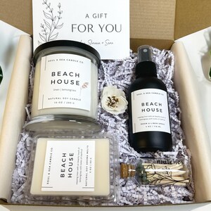 May include: A gift box filled with a white candle, room spray, and matches. The candle and room spray are labeled "Beach House" and have a natural soy candle scent. The gift box is decorated with white shredded paper and a small white flower.