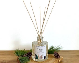 Puede incluir: Un frasco difusor de vidrio transparente con varillas de madera, etiquetado como "Fraser Fir". La etiqueta presenta un diseño de bosque con árboles verdes, azules y blancos. El difusor se encuentra sobre una rodaja de madera con agujas de pino y una piña.