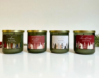 Puede incluir: Se muestran cuatro velas de vidrio verde con tapas doradas. Cada vela tiene una etiqueta con texto y un diseño de invierno. Las etiquetas dicen "Cedar Balsam", "Winter Berry", "Winter Woods" y "Christmas Morning".