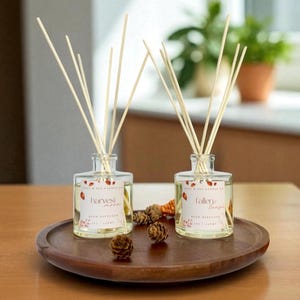 May include: Two clear glass reed diffusers with wooden reeds, labeled "harvest" and "fallen" on a wooden tray. The diffusers have a light-colored liquid and are decorated with fall-themed leaves. Small pinecones are scattered around the diffusers.
