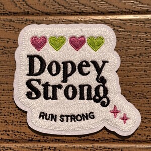 May include: White embroidered patch with the text "Dopey Strong" and the words "Run Strong" below. The patch has four hearts in a row, two pink and two green, and two pink stars.