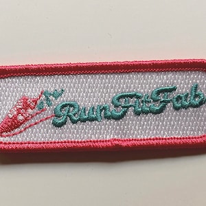 May include: Embroidered patch with a rectangular shape and a pink border. The patch features the words "Run Fit Fab" in teal script, along with a stylized carrot design. The background is white with a textured pattern.