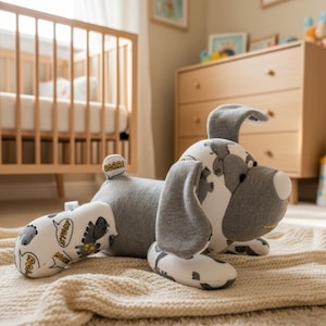 May include: A plush dog toy featuring a gray and white fabric with a repeating pattern of cartoon dinosaurs and speech bubbles containing the text ROAR and STOMP. The toy has a solid gray midsection, floppy ears, and a white nose detail.