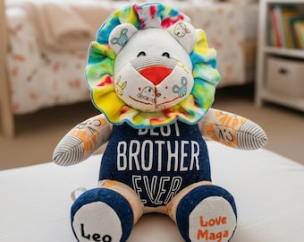 Puede incluir: Un león de peluche con melena tie-dye, cara blanca con pequeñas ilustraciones de animales y torso azul oscuro con el texto blanco BEST BROTHER EVER. Los pies están bordados con el texto negro Leo y el texto naranja Love Maga.