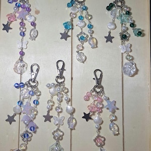 May include: A collection of keychains with silver clasps, featuring beaded strands with seashell, star, and butterfly charms. The keychains showcase a variety of colors, including pink, blue, green, and iridescent beads, creating a coastal theme.