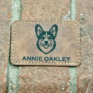 Könnte beinhalten: Ein rechteckiger, brauner Lederaufnäher mit einer genähten Umrandung. Der Aufnäher zeigt eine grüne Illustration eines Hundegesichts und den Text "ANNIE OAKLEY" in dunkler Schrift. Der Aufnäher befindet sich vor einem Ziegelhintergrund.