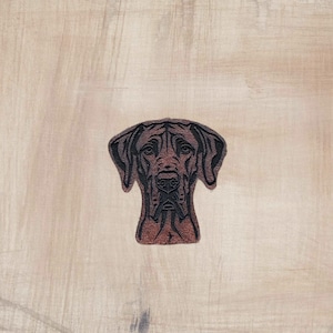 May include: A brown and black dog patch with a detailed design. The patch features a dog's head and shoulders, with a focus on the face and ears. The background is a light brown, textured surface.