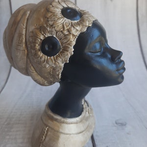 May include: A black bust of a woman with a gold-coloured head wrap and collar. The head wrap features two large, dark-centred flower designs. The bust is set against a light-coloured wooden background.