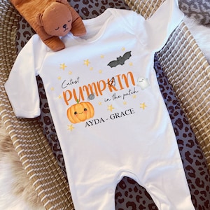 May include: White baby onesie with long sleeves, featuring a Halloween-themed design. The onesie has a smiling pumpkin, a bat, a spider, and the text "Cutest Pumpkin in the patch". The name "AYDA - GRACE" is also printed on the onesie.
