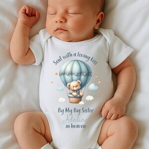 May include: A white baby onesie with a blue hot air balloon design, a teddy bear, and the text "Sent with a loving kiss By My Big Sister Adeline in heaven". The onesie is short-sleeved.