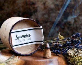 Könnte beinhalten: Ein brauner Räucherkegel mit aufsteigendem Rauch, neben einem Behälter mit der Aufschrift "Lavender Frankincense + Cedar". Getrockneter Lavendel und Moos befinden sich im Hintergrund. Der Weihrauch liegt auf einer Holzoberfläche.