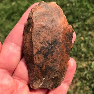May include: A roughly shaped, reddish-brown stone tool with dark brown and black markings. The stone has a textured surface and appears to be hand-chipped, with a sharp edge. The object is held in a hand against a blurred green background.