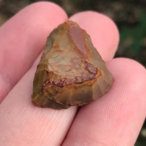 May include: A small, triangular, stone arrowhead with a mottled appearance. The arrowhead features shades of brown, tan, and reddish-brown, with a slightly translucent quality. The edges are sharp and well-defined, suggesting it was crafted for hunting or warfare.