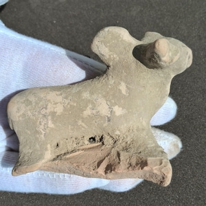 May include: A small, weathered, tan-colored clay figurine of an animal, possibly a dog or a deer. The figurine has a rough texture and appears to be an ancient artifact. The animal has a small head, ears, and a body with a tail.
