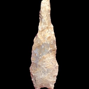 May include: A light-colored, stone tool with a pointed tip and a rough, textured surface. The artifact is set against a black background, highlighting its shape and details. The tool appears to be hand-crafted.