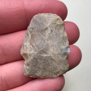 May include: A light brown, irregularly shaped stone tool with a pointed tip. The tool is held in a hand, but the hand is not visible in the image.