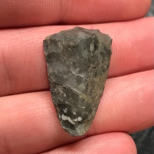 May include: A gray and white arrowhead made of flint. The arrowhead is pointed and has a sharp edge.