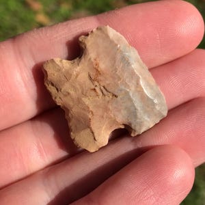 May include: A small, tan and gray arrowhead with a chipped edge. The arrowhead is held in a hand.