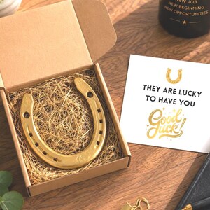 Könnte beinhalten: Ein goldfarbenes Metallhufeisen in einer braunen Pappschachtel mit strohähnlichem Füllmaterial. Neben der Schachtel liegt eine weiße quadratische Karte mit dem Text "THEY ARE LUCKY TO HAVE YOU" und "Good Luck!" in Gold und Schwarz, ein schwarzer Becher und ein Stift.