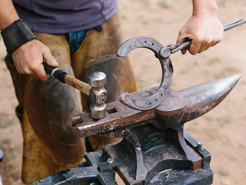 May include: A blacksmith forges a horseshoe on an anvil. The horseshoe is held with tongs while being hammered. The anvil is dark metal, and the blacksmith wears leather protective gear.