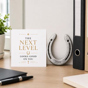 May include: A square white greeting card with gold and black text reading THE NEXT LEVEL LOOKS GOOD ON YOU, featuring a small horseshoe icon. Beside it sits a silver-toned metal horseshoe with the word Boss engraved at the bottom center.