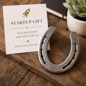Peut inclure: Un fer à cheval argenté est posé sur une surface en bois, à côté d'une carte avec l'inscription « STARTUP GIFT ». La carte contient également le texte « Every great journey starts with a bold step. BELIEVE. BUILD. SUCCEED. »