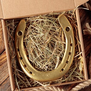 May include: A gold-colored horseshoe rests in a cardboard box filled with hay. The horseshoe is U-shaped with nail holes and is presented on a wooden surface with a rope border. The image has a rustic, Western theme.