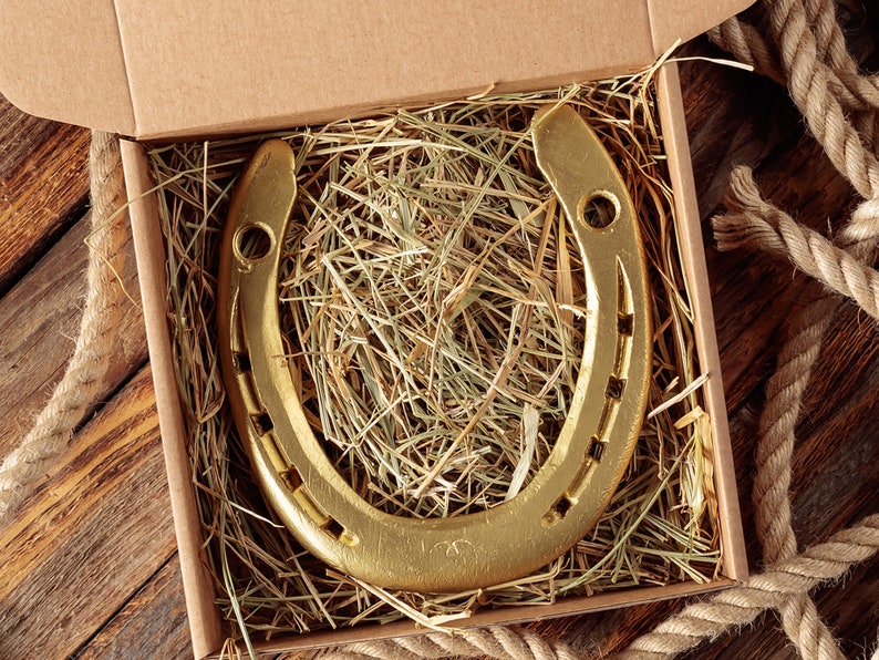 May include: A gold-colored horseshoe rests in a cardboard box filled with hay. The horseshoe is U-shaped with nail holes and is presented on a wooden surface with a rope border. The image has a rustic, Western theme.