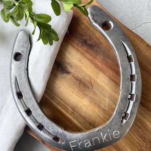 Może przedstawiać: Srebrna podkowa z wygrawerowanym imieniem "Frankie". Leży na drewnianej desce z białym płótnem i zielonymi liśćmi. Podkowa, symbol szczęścia, to tradycyjny prezent lub przedmiot dekoracyjny.