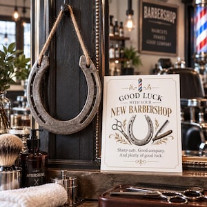 Könnte beinhalten: Ein Metallhufeisen, das an einem Juteseil hängt, neben einem weißen Schild mit der Aufschrift Good Luck With Your New Barbershop, illustriert mit einem Barber-Pole, einer Schere, einem Rasiermesser und dem Text Sharp cuts. Good company.