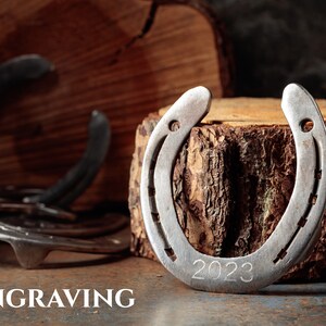 May include: A silver horseshoe with the engraved year "2023" is displayed on a wooden surface. The word "ENGRAVING" is at the bottom of the image. Additional horseshoes are visible in the background, suggesting a theme of luck or equestrianism.