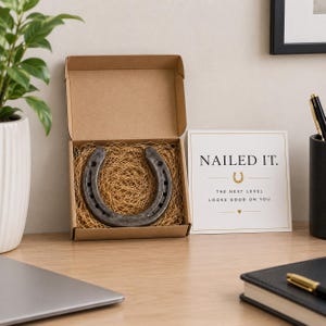 Puede incluir: Una herradura de metal gris oscuro colocada en una caja de regalo de cartón marrón rellena con papel triturado beige. Junto a la caja hay una tarjeta blanca con texto dorado que dice NAILED IT. THE NEXT LEVEL LOOKS GOOD ON YOU con un icono.
