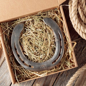 Rustic used horseshoe in a kraft gift box filled with natural hay, placed on a wooden surface with thick rope beside it — authentic handmade good luck charm.