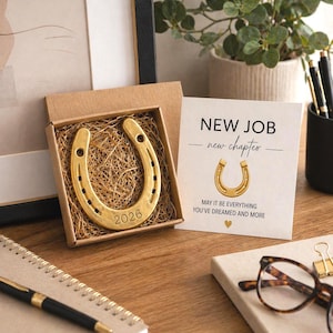May include: A gold-colored horseshoe with the year 2026 engraved, presented in a small cardboard box filled with straw. A card next to it reads "NEW JOB - new chapter" with a horseshoe graphic and a message. The scene is set on a wooden surface.