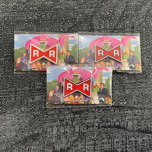 May include: Three collectible pins featuring a pink monster, a red and white logo with the letters "R", and a background of characters. The pins are in clear packaging, with the words "COLLECTIBLE PIN" visible.