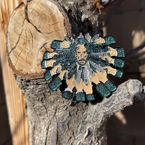 May include: A detailed enamel pin featuring a stylized portrait of a man surrounded by multiple hands holding guns. The pin has a glittery green and gold color scheme, with a black suit and tie. The design is intricate and eye-catching.