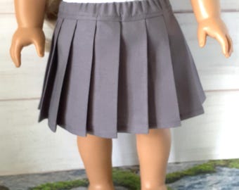 May include: A gray pleated skirt for a doll. The skirt is made of a solid gray fabric and has a pleated design. The skirt is paired with a white top. The doll is standing on a stone surface.