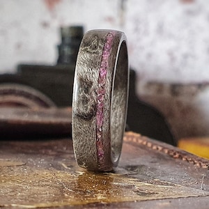May include: A dark grey wooden ring with a thin band of pink stones embedded in the wood.