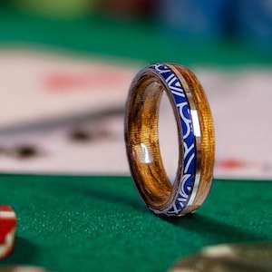 Op de afbeelding: Een houten ring met een blauw en zilver inlegontwerp. De ring is gemaakt van lichtbruin hout en heeft een decoratief patroon rond de band. De ring wordt gepresenteerd op een groene ondergrond met pokerchips en speelkaarten op de achtergrond.