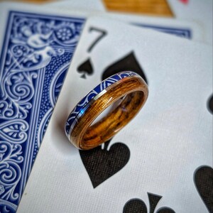 Op de afbeelding: Een houten ring met een blauwe en zilveren inleg, rustend op speelkaarten. De ring heeft een natuurlijke houtnerf en een decoratieve blauwe band. De speelkaarten tonen een blauw patroon op de achterkant en de zeven schoppen.
