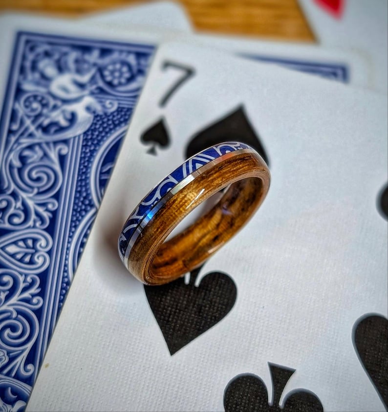 Op de afbeelding: Een houten ring met een blauwe en zilveren inleg, rustend op speelkaarten. De ring heeft een natuurlijke houtnerf en een decoratieve blauwe band. De speelkaarten tonen een blauw patroon op de achterkant en de zeven schoppen.