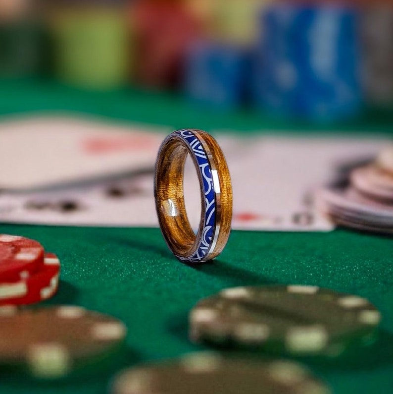 Op de afbeelding: Een houten ring met een blauw en zilver inlegontwerp. De ring is gemaakt van lichtbruin hout en heeft een decoratief patroon rond de band. De ring wordt gepresenteerd op een groene ondergrond met pokerchips en speelkaarten op de achtergrond.