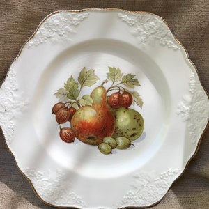 May include: White ceramic plate with a gold rim and a raised floral design. The center of the plate features a painted image of pears and gooseberries.